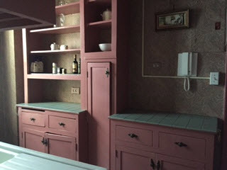 Dark rose painted cabinets and open shelves, green ceramic tile counters, pale paisley wallpaper in the kitchen